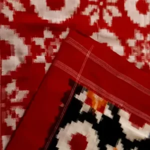 Close folded view of a red Double Ikat pure cotton saree showcasing traditional white geometric motifs on the body, paired with a contrasting multicolor black border featuring intricate Ikat patterns and fine red stripe detailing.