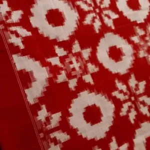 Close view of a red Double Ikat pure cotton saree showcasing traditional white geometric and floral-style motifs with a symmetrical woven pattern, captured on a smooth fabric surface.