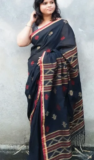 Woman draped in a black handloom cotton Jamdani saree with red and gold patterns, standing against a plain background.