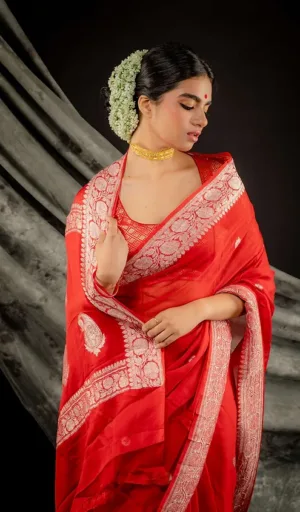 A woman wearing a red saree with intricate silver embroidery, adorned with a floral hairpiece and jewelry, posing elegantly.
