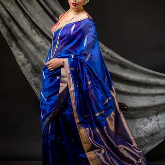 Handloom-Pure-Silk-Chanderi-5 Woman wearing a royal blue pure silk Chanderi handloom saree with silver zari border and motifs, posing against a draped backdrop; elegant festive silk saree look.
