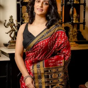 Woman wearing a red handloom silk saree with intricate patterns and a black blouse, standing in an indoor setting.