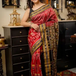 Handloom pure silk Odisha Ikat saree in rich red, featuring intricate temple borders, multicolour geometric motifs, and a traditional black-and-gold woven pallu, draped by a woman in a black blouse inside a heritage décor setting.