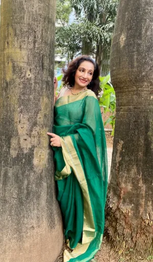 Woman wearing a green saree with gold accents, posing between two large trees in an outdoor setting.