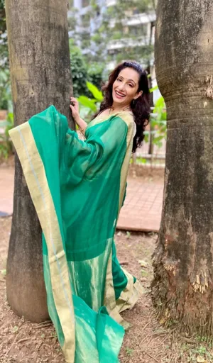 Woman wearing a green and gold saree, smiling while standing between two trees in an outdoor setting.