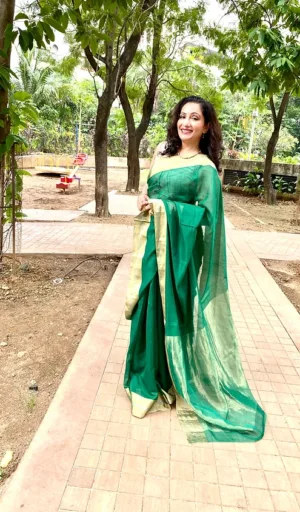 Woman wearing a green handloom cotton silk Chanderi saree with a gold border, standing outdoors with trees in the background.