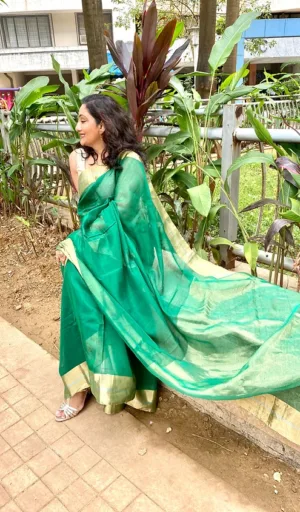 Woman wearing a green handloom cotton-silk Chanderi saree with a flowing pallu, standing outdoors with greenery in the background.