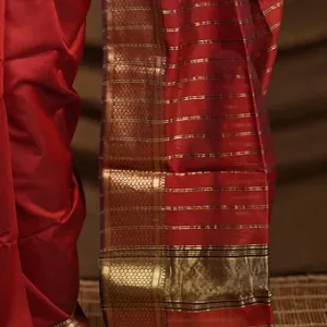 Woman wearing a maroon handloom cotton silk Maheshwari saree with gold zari stripes and rich border, standing in a traditional setup; elegant festive Maheshwari saree look.
