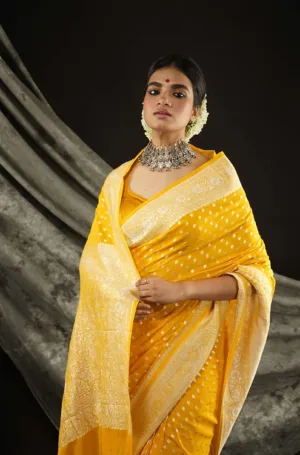 A woman wearing a yellow saree with intricate patterns and a matching necklace, posing elegantly against a dark background.