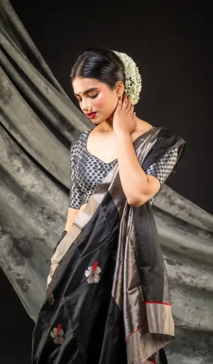 A woman wearing a black saree with floral embroidery and a patterned blouse, adorned with a floral hair accessory.