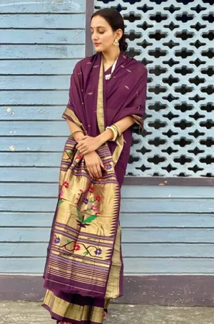 A woman wearing a purple handloom pure cotton Paithani saree with intricate gold detailing and floral embroidery, standing against a decorative wall.
