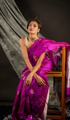 A woman wearing a vibrant pink handloom silk Chanderi saree, seated on a wooden chair with a graceful pose.