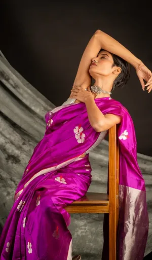 Model wearing a vibrant purple handloom silk Chanderi saree with floral patterns, seated gracefully on a wooden chair.