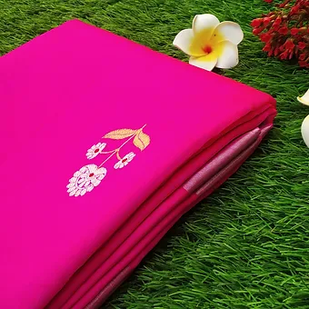 Pink Pure Silk Chanderi Saree with Floral Butta & Silver Zari Border Folded pink pure silk Chanderi saree featuring delicate white floral butta embroidery near the pallu and a soft silver zari border, placed on green grass with decorative flowers.