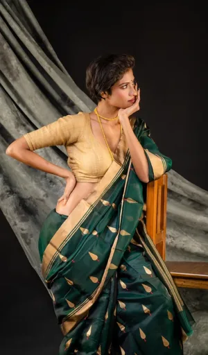 Woman wearing a green handloom saree with gold leaf patterns and a beige blouse, seated in a thoughtful pose.