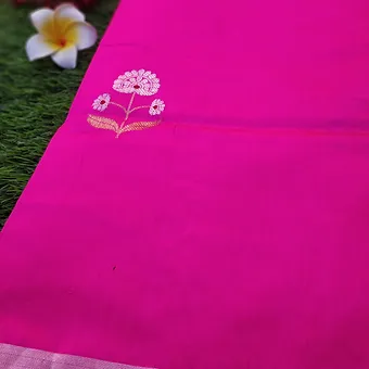 Pink Pure Silk Chanderi Saree with White Floral Butta & Silver Border Close view of a bright pink pure silk Chanderi saree featuring delicate white floral butta embroidery near the pallu and a subtle silver zari border, displayed on a grass background with a plumeria flower.
