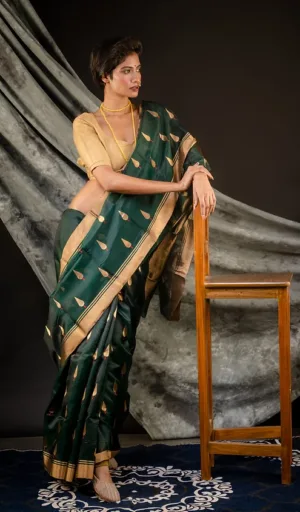 Model wearing a green handloom silk saree with gold motifs, paired with a beige blouse, standing beside a wooden chair.