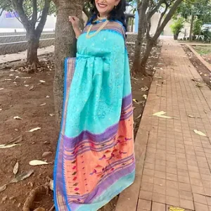 A woman wearing a turquoise and orange handloom silk saree with intricate patterns, standing beside a tree in a natural setting.