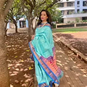 Woman wearing a light blue handloom silk saree with a colorful border, standing outdoors among trees.