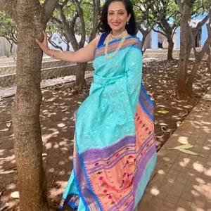 Woman wearing a light blue saree with intricate designs and a contrasting orange border, posing outdoors among trees.
