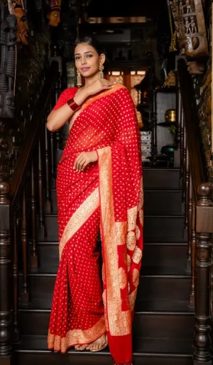 Woman wearing a red saree with gold polka dots and intricate border, standing on a staircase with traditional decor.