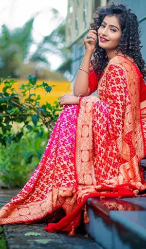 Model wearing a Handwoven Pure Chiffon Banarasi saree in shades of red and pink, adorned with intricate gold zari motifs. The saree is elegantly draped, showcasing its traditional charm with a beautifully detailed border