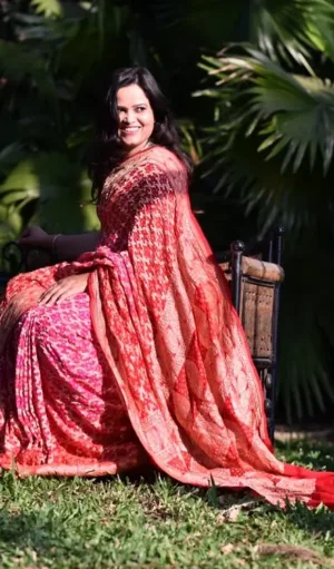 Model wearing a Handwoven Pure Chiffon Banarasi saree in shades of coral and pink, featuring intricate gold zari patterns. The saree is elegantly draped with a rich, flowing pallu, offering a traditional yet modern look against a natural backdrop.