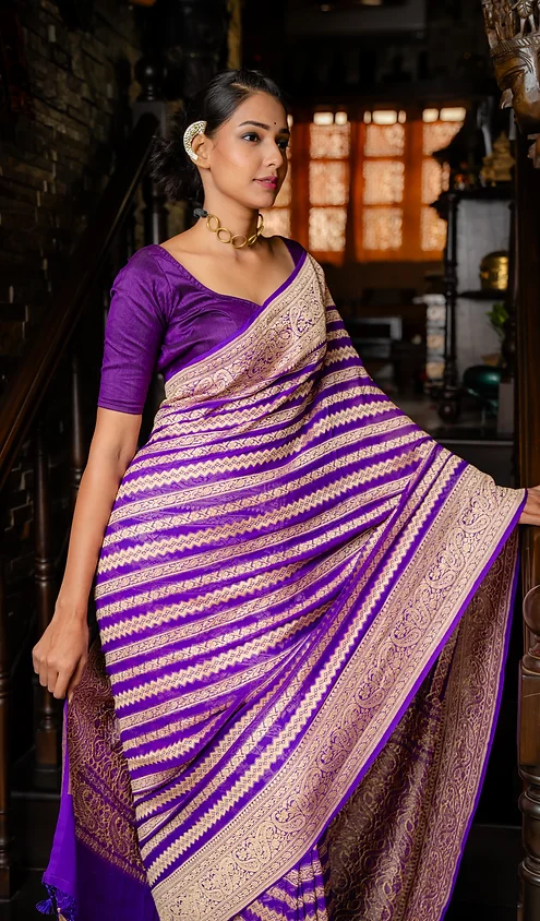 Handwoven-Pure-Khaddi-Georgette-Banarasi-1 Woman wearing a purple saree with gold and silver stripes, standing on a staircase with a traditional setting.
