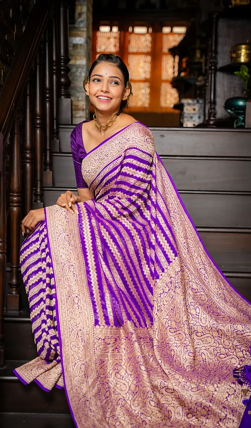 Handwoven-Pure-Khaddi-Georgette-Banarasi-3 Woman wearing a purple and beige handwoven saree with intricate patterns, seated on a staircase with a smile.