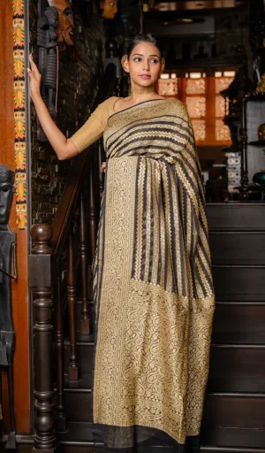 A woman wearing a handwoven saree with a black and gold design, standing on a staircase with decorative elements in the background.