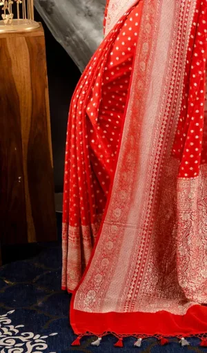 Close-up view of Handwoven Pure Khadi Georgette Banarasi saree in red, featuring white polka dots and a beautiful gold zari border with floral patterns. The saree is finished with a red tassel edge, adding a traditional and elegant touch.