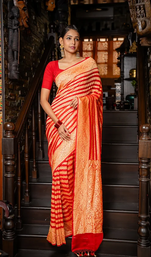 Handwoven-Pure-Khaddi-Georgette-banarasi-2 A woman wearing a red and orange striped saree with intricate golden patterns, standing on a staircase.
