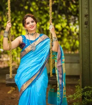 Woman wearing a blue cotton Paithani saree with intricate designs, seated on a swing, adorned with jewelry and a traditional hairstyle.
