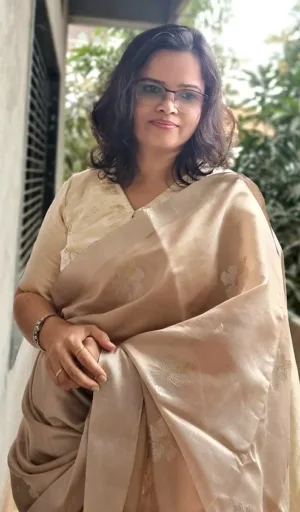 Woman wearing a beige handloom silk Chanderi saree with floral patterns, standing with a gentle smile and holding the saree drape.