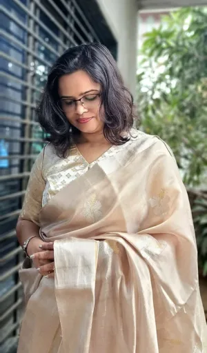 Woman wearing a beige handloom silk Chanderi saree with intricate gold embroidery, looking down thoughtfully with soft natural light.
