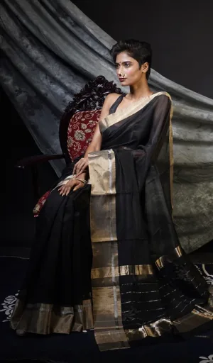 A woman wearing a black handloom pure silk Chanderi saree with gold accents, seated elegantly on a chair.