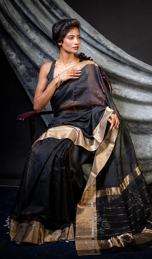 handloom-pure-silk-chenderi-2 A woman seated in a black saree with gold accents, showcasing traditional attire with a modern touch, styled elegantly against a dark backdrop.