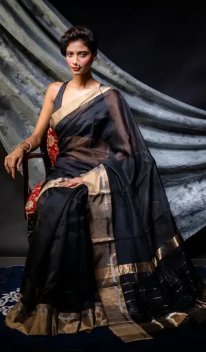 Model wearing a black handloom pure silk Chanderi saree with gold accents, seated on a chair against a dark backdrop.