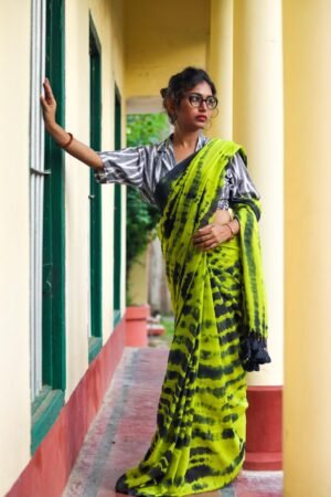 A woman wearing a green mul cotton tie-dye saree with black patterns, standing against a wall with her hand resting on it.