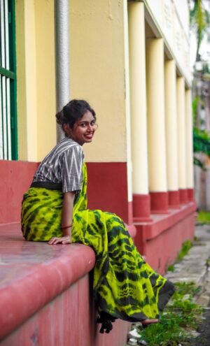 Green mul cotton tie and dye saree worn by a woman sitting on a ledge, showcasing intricate patterns and vibrant colors.
