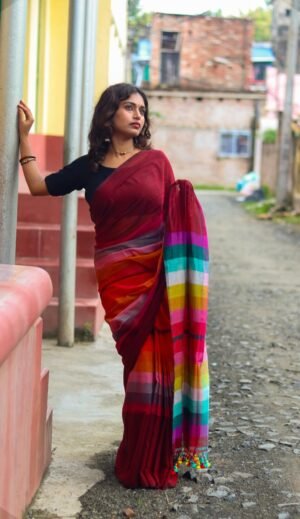 Handwoven mul cotton saree in red with multicolored striped pallu, worn by a woman standing against a wall.