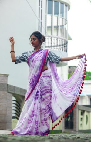 A woman wearing a purple mul cotton tie-dye saree with a contrasting border and tassels, posed gracefully outdoors.