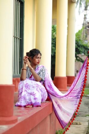 A woman wearing a purple tie-and-dye cotton saree with a silver blouse, seated on a ledge with a flowing pallu adorned with red tassels.