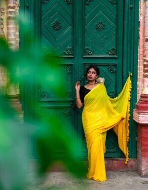 Woman wearing a yellow handwoven mul cotton saree with a black blouse, standing beside a green door.